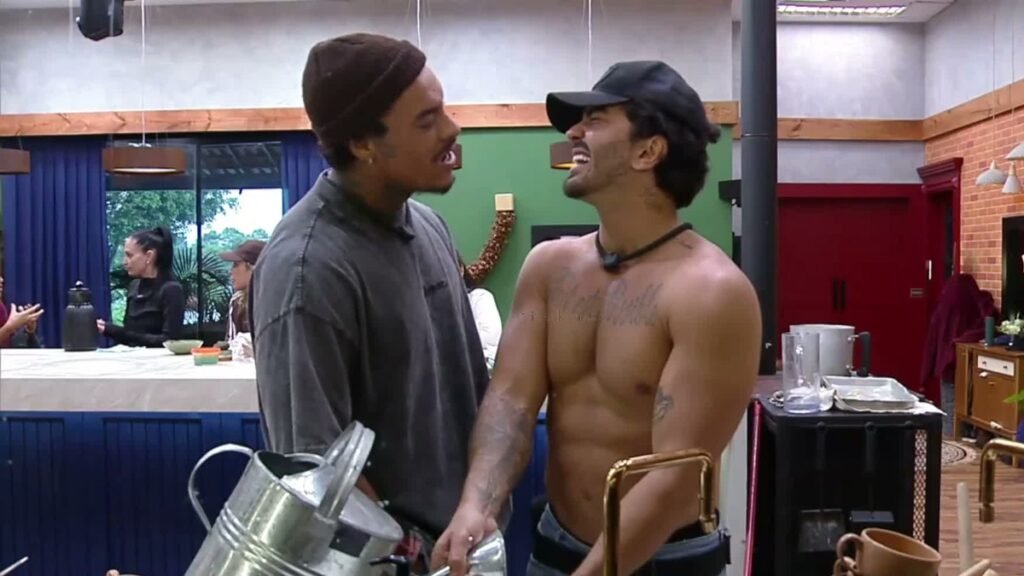 A Fazenda 18: Murilo confronta Alfredo na pia, cobra a promessa do chicote e desafia: “Pode vir!” Murilo Dias, de camiseta cinza e gorro, cara a cara com Alfredo Amaral, sem camisa e de boné, junto à pia da cozinha da sede de A Fazenda 18